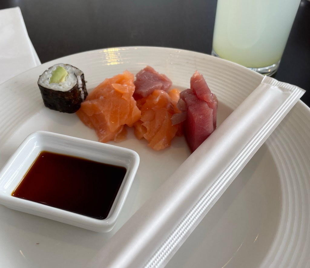 A plate holds one piece of a California roll, two pieces of salmon sashimi, two pieces of tuna sashimi, a small bowl of soy sauce, and a set of chopsticks. 