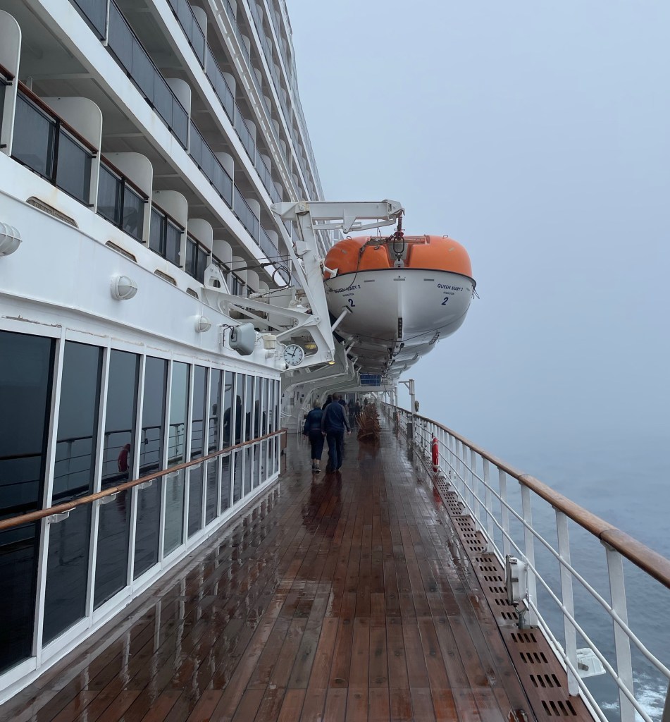 A view down the lengthy teak promenade deck on Mary 2 shows a few passengers walking despite the rainy weather. A wall of windows is next to the promenade as five decks of cabins with private balconies extend the view upward. 