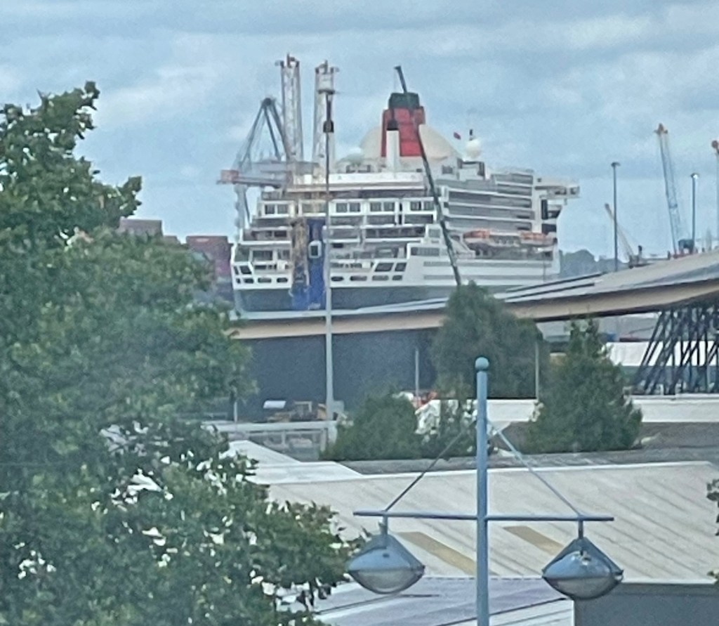 The liner Queen Mary 2 as viewed from a closed window of a building on land, showing her aft decks, most of her starboard side, and bright funnel. 