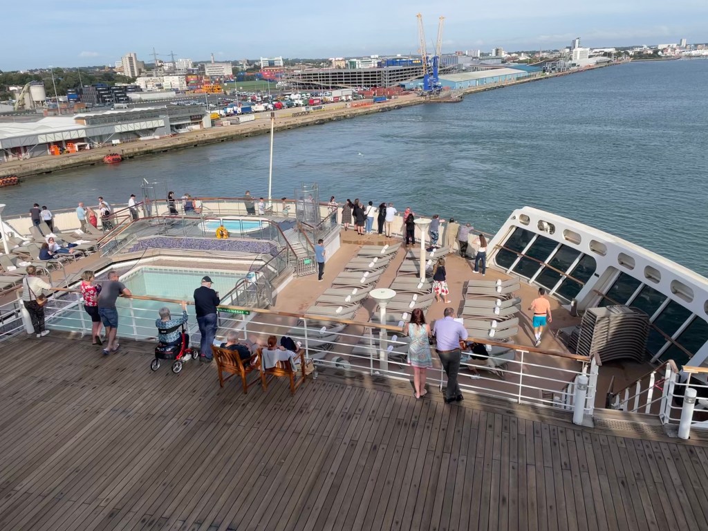 Looking aft from an upper deck as Queen Mary 2 leaves the terminal. Two more decks cascade below, one with a swimming pool but no swimmers yet, and both lined with a couple of dozen passengers enjoying the view of the river over the stern. 