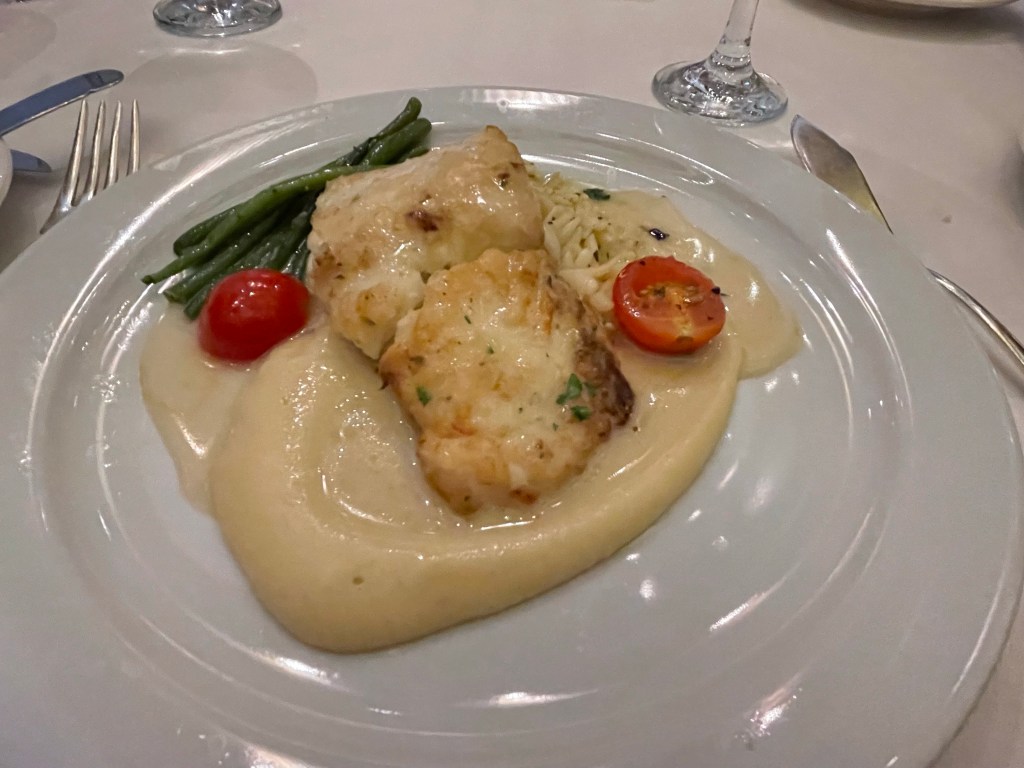 Dinner plate shows a browned filet of cod about one quarter the size of the plate atop a sauce of puréed yellow vegetable with a cherry tomato cut into halves and about 6 asparagus spears. 