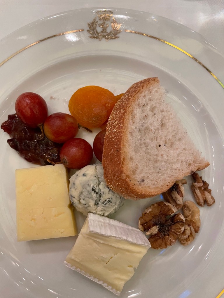 A gold trimmed dessert plate with the Cunard logo on top in gold holds a chunk of French bread, an ounce each of a white cheddar, Brie, and blue cheese, walnuts, and five pieces of dried fruit. 