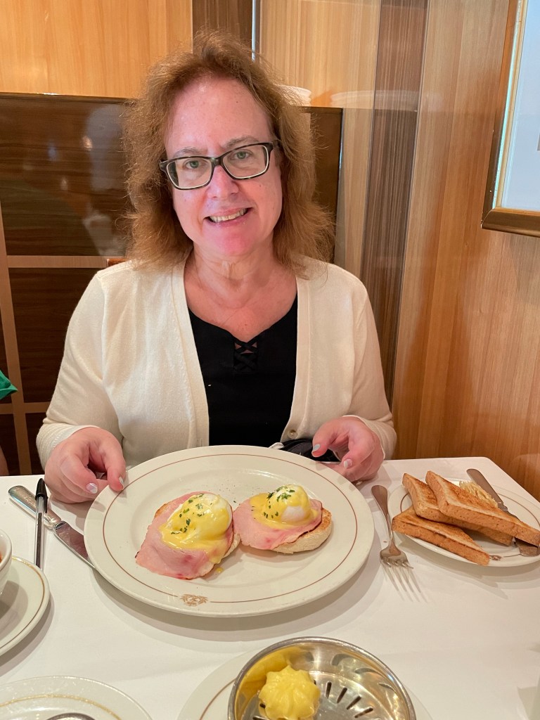 Lisa is seated at a restaurant table behind a plate of Eggs Benedict on 2 English muffin halves. 