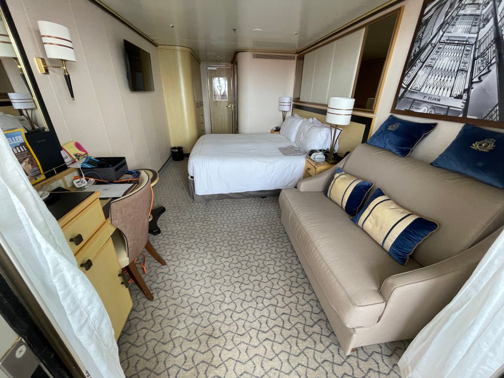 On the left is a desk, chair and mirror. On right is a sofa with four throw pillows under a black and white art deco architectural print. The double bed is further back, its headboard against the wall on the right.