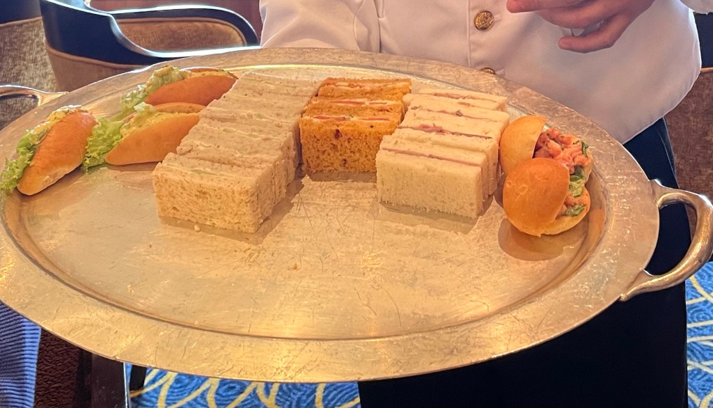 Small sandwiches lined up neatly in five rows by type, some on crustless bread, some on rolls, on a silver platter held by a waiter in formal attire