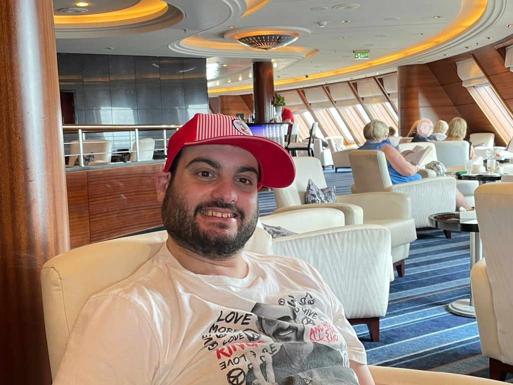 Young man in his 20s smiles while sitting in the front of the lounge, the port side visible behind him. Windows tilt inward and everyone in the photo is seated in a large upholstered white chair and dressed casually for the day. 