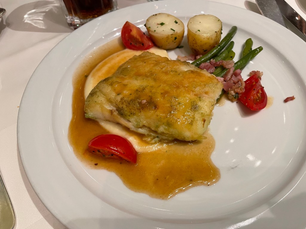 A fish filet approximately one third the size of the dinner plate sits atop a purée alongside two small potato halves tomato green beans and a red wine sauce