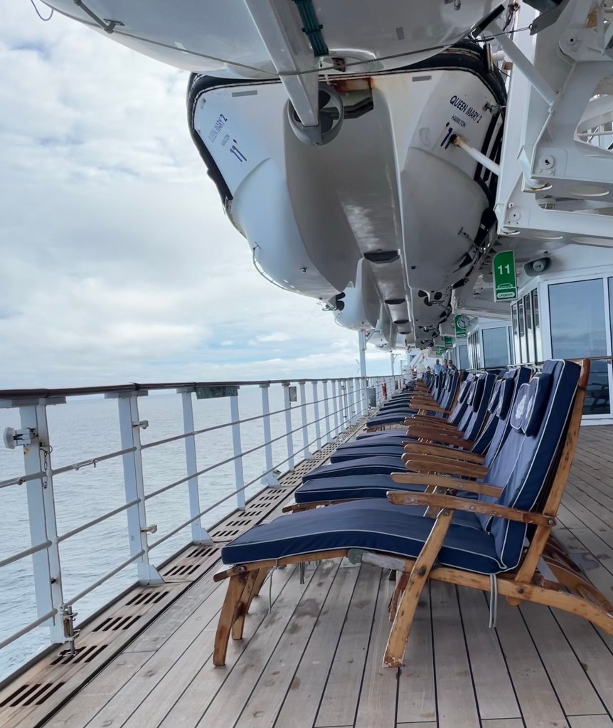 A row of wood deck chairs with cushioned seats and backs line the promenade deck of a ship under the lifeboats and along the rail overlooking the ocean.