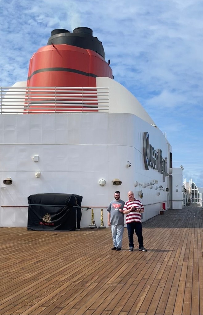 Neil and Marty appear tiny below the massive Cunard funnel that rises from one deck above the wide open teak sports deck they are standing on.