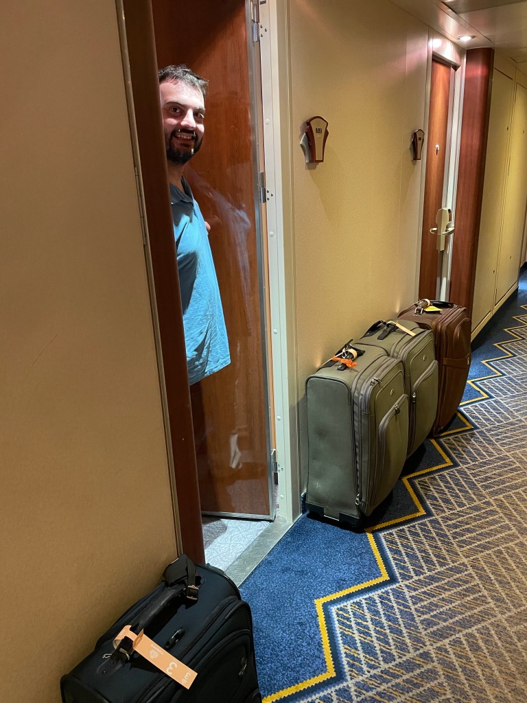 Marty stands in the doorway of a cabin into a hallway where several suitcases line the walls. 
