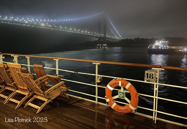 From the deck of Queen Mary 2, three wood deck chairs and a life ring are in the foreground as the ship passes under the Verrazano Bridge in New York City.
