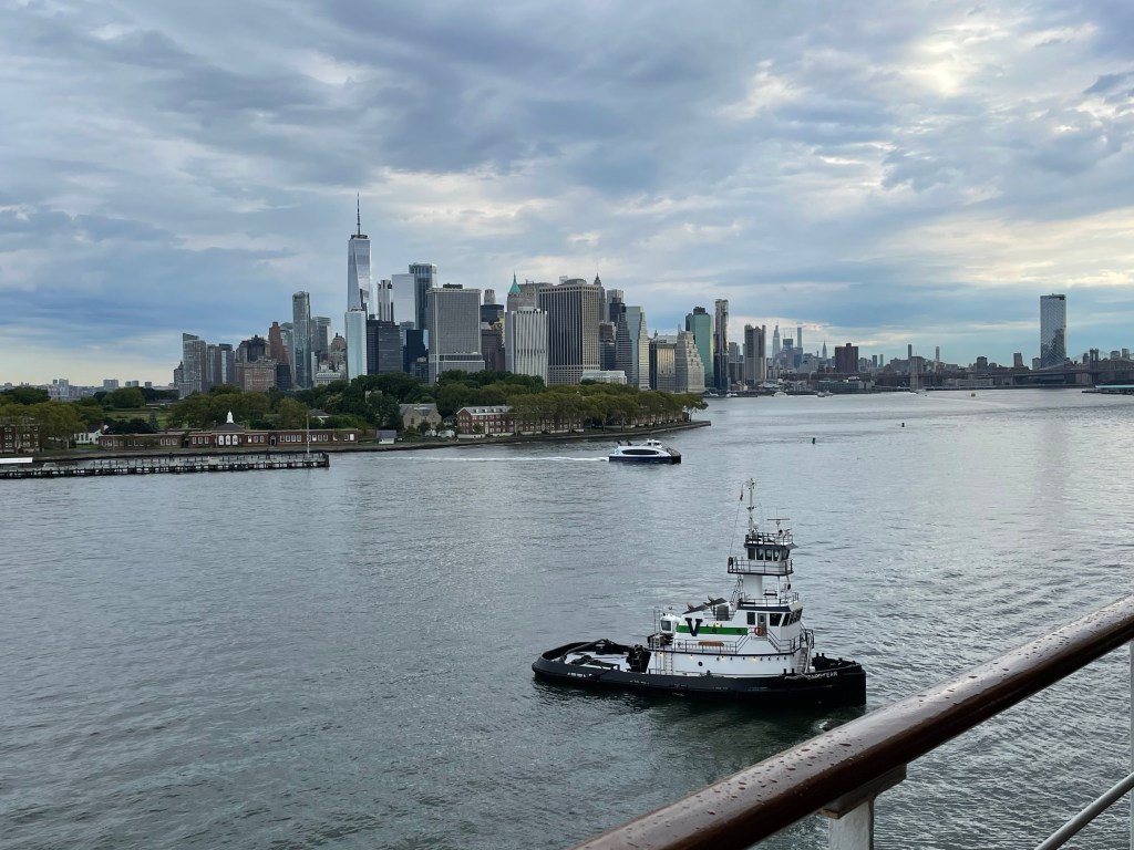 A tug is alongside Queen Mary 2 as viewed from the liner, with the skyline of lower Manhattan in the background 