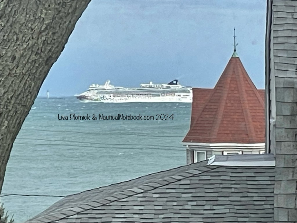 A midsize cruise ship is off in the distance in profile the camera placing it between a nearby tree and nearby Victorian house. The ship appears wavy due to the glass window the photographer was standing behind. 