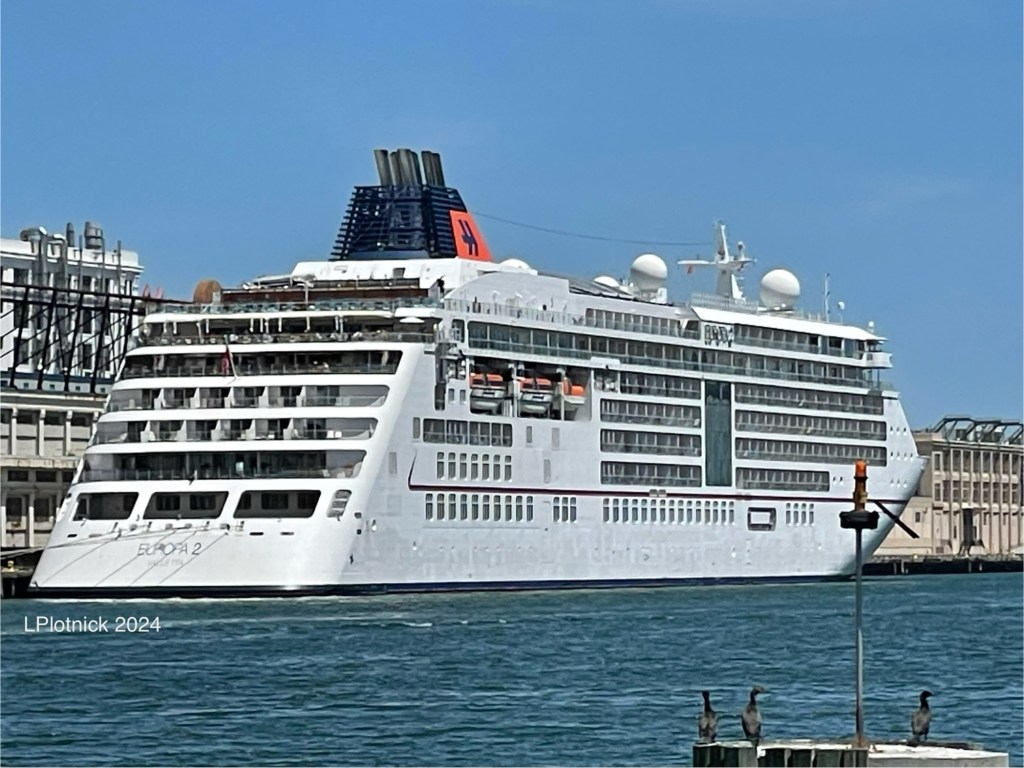 The all white ship Europa 2 in aft and starboard view, showing 7 decks on an outward sloping stern, rows of inset balcony cabins and 3 lifeboats starboard, and a bright orange funnel. 