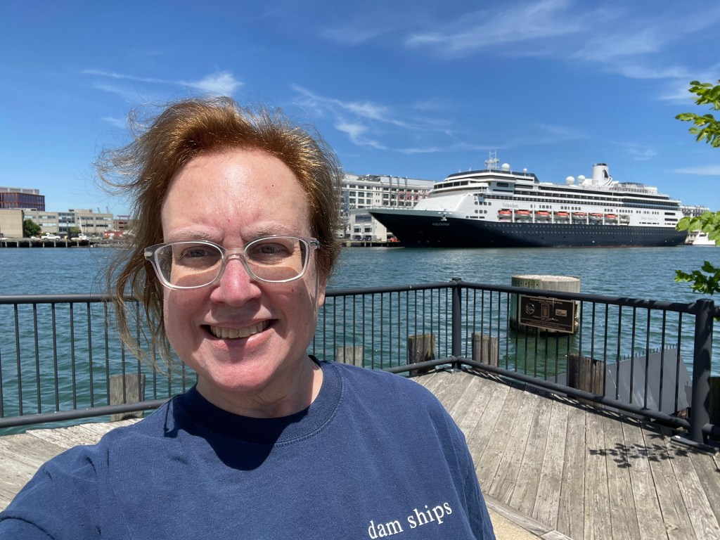 A selfie of Lisa with the dark blue hulled Volendam in port side profile. Lisa’s t shirt reads like d a m ships. The ship has classic lines, with picture window cabins below a promenade, large windows near the overhanging lifeboats, and 2 levels of balcony cabins near the top of the ship.  