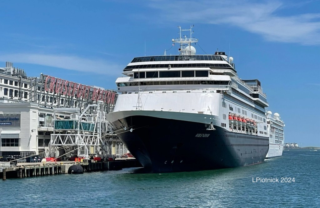 A glass enclosed gangway leads from the Boston cruise terminal to the forward facing holland America ship Volendam as the small all white cruise ship Europa 2 is somewhat visible behind Volendam. 