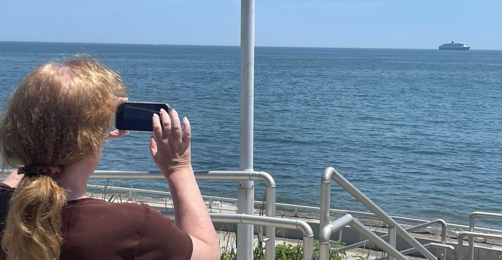 Lisa uses a smartphone to photograph Queen Mary 2, which is off on the near horizon. Lisa is seen from behind, her light brown hair pulled back into a ponytail. 