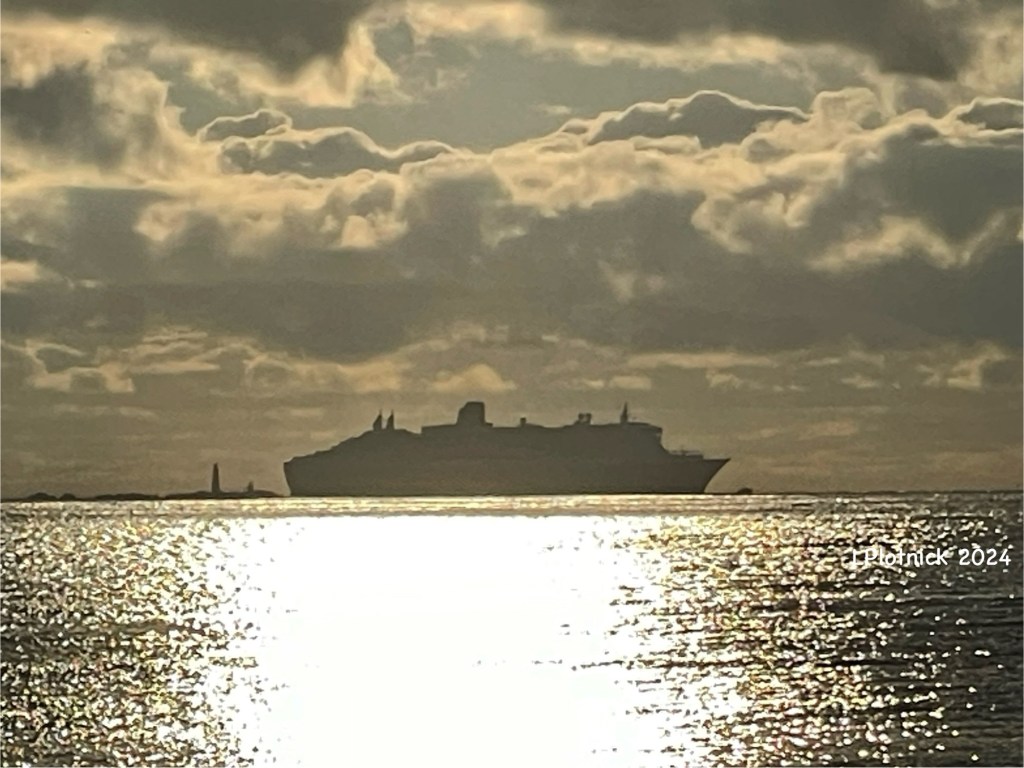 Queen Mary 2 in profile soon after sunrise, the sun’s rays reflecting on the ocean between the ship and the shore. 