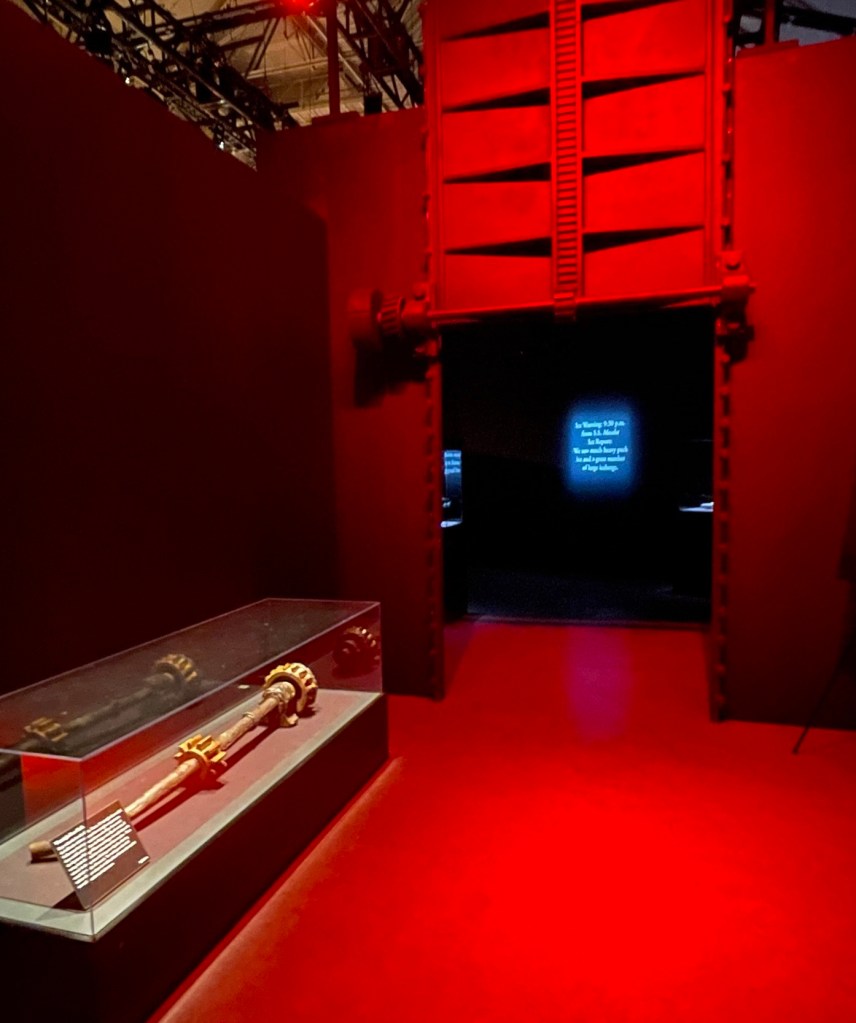 Bright red walls and floor in a room containing a gold shaft about 6 feet long laying in a glass case and a wall opening with a replica of a watertight door hanging above it. 