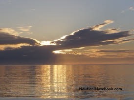 The sun just above the horizon and behind wispy clouds forms a golden reflection on a calm sea. 