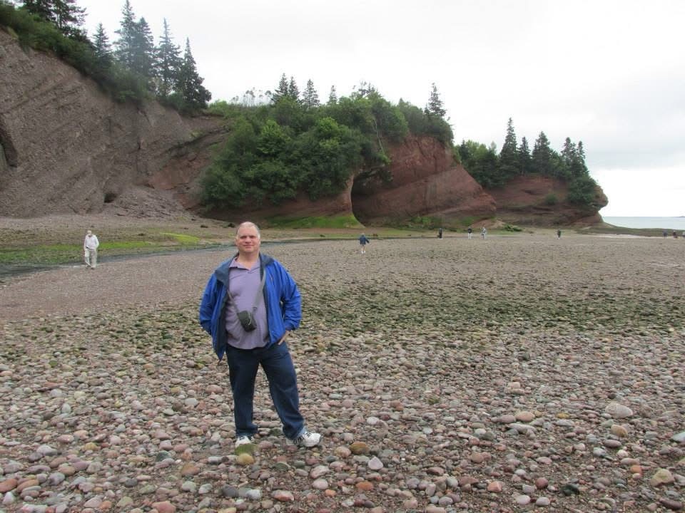 The author’s husband stands on a rock covered beach at low tide on a cool and overcast day. In the background close to the waters edge is a grouping of mud covered caves. 