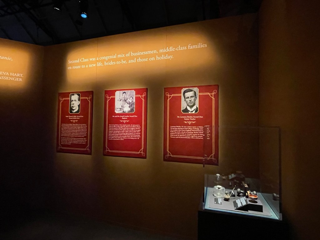 Along a wall under these words Second Class was a congenial mix of businessmen, middle-class families en route to a new life, brides-to-be, and those on holiday are bio panels of an older man, a young family, and a younger man. In the foreground is a plexiglass case containing 4 small items including a doorknob and bowl. 