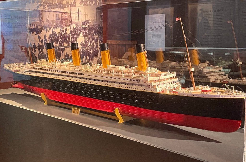 Inside a glass case a detailed model of Titanic features the ship’s four yellow and black funnels, a red and black hull, and intricate details such as windows, portholes, wood decking, and mechanical equipment. 