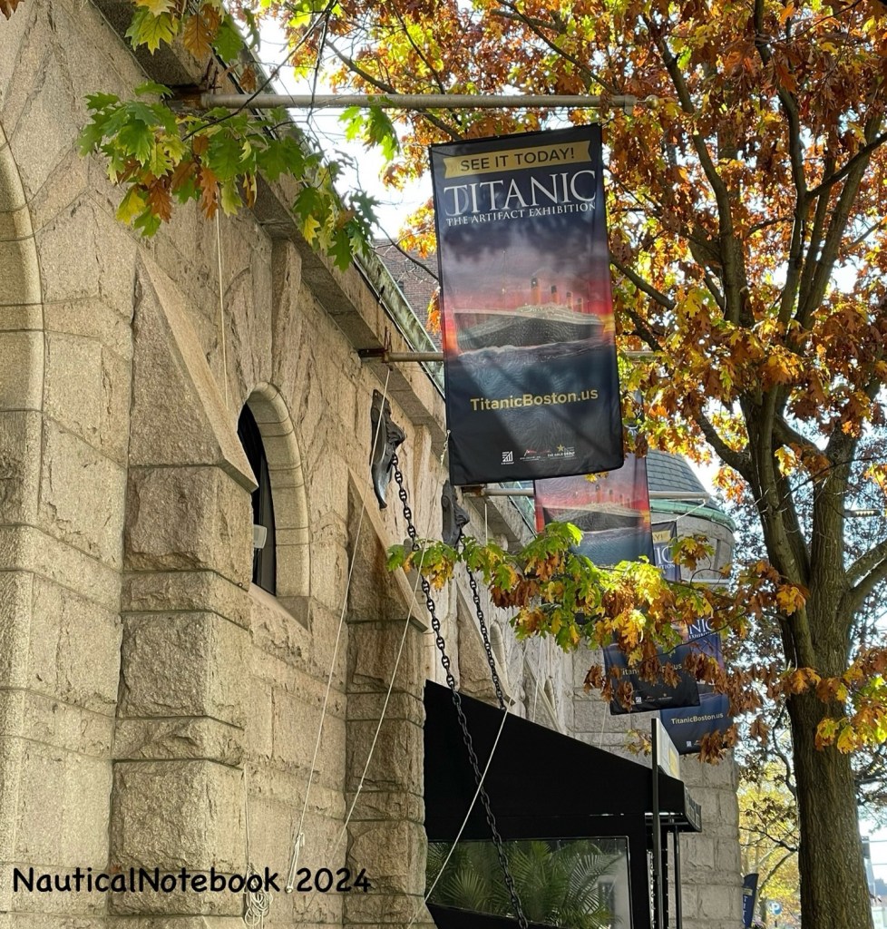 Banner hangs from a flagpole above an arched window and square entryway of a stone building. It reads see it today titanic the artifact exhibition website titanic Boston dot U S. 