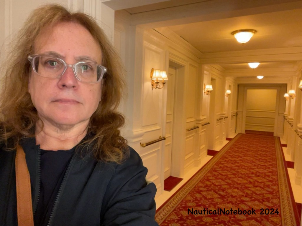 Lisa stands at the entrance to the first class cabin corridor which features white walls and ceiling, a red carpet with gold trim, 3 flush mounted ceiling lights, 6 doors each flanked by a wall sconces with two lights and shades, and wood rails at hip height between the cabin doors. 