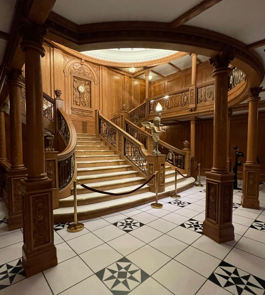 Replica of Titanic’s grand staircase includes the lobby on the lower landing with its geometric tiles and floor to ceiling wooden columns, and intricate detail of the stair railings, cherub statue, and on the upper landing the honor and glory carving. The famous glass dome is just out of range. 
