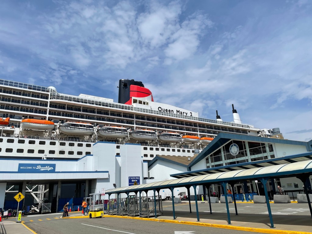 The modern day ocean liner Queen Mary 2 rises above the Brooklyn cruise terminal, a set of 2 squat buildings, one with a steepled roof above the entry, and a vehicle drop off point with covered walkway, under a blue sky with wispy white clouds. 