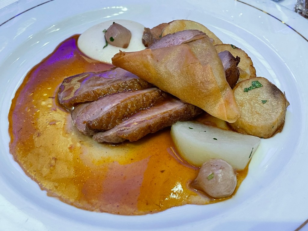 3 slices of skin on roasted duck topped with a puff pastry containing onion, a small scoop of a white puree, potatoes, and a brown sauce. 