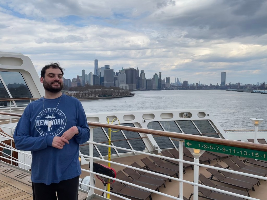 A young Caucasian man with dark hair and beard poses  on a deck overlooking an aft deck with many empty loungers and in the distance the skyline of New York City. He is wearing a long sleeved graphic shirt that reads New York the city that never sleeps. 