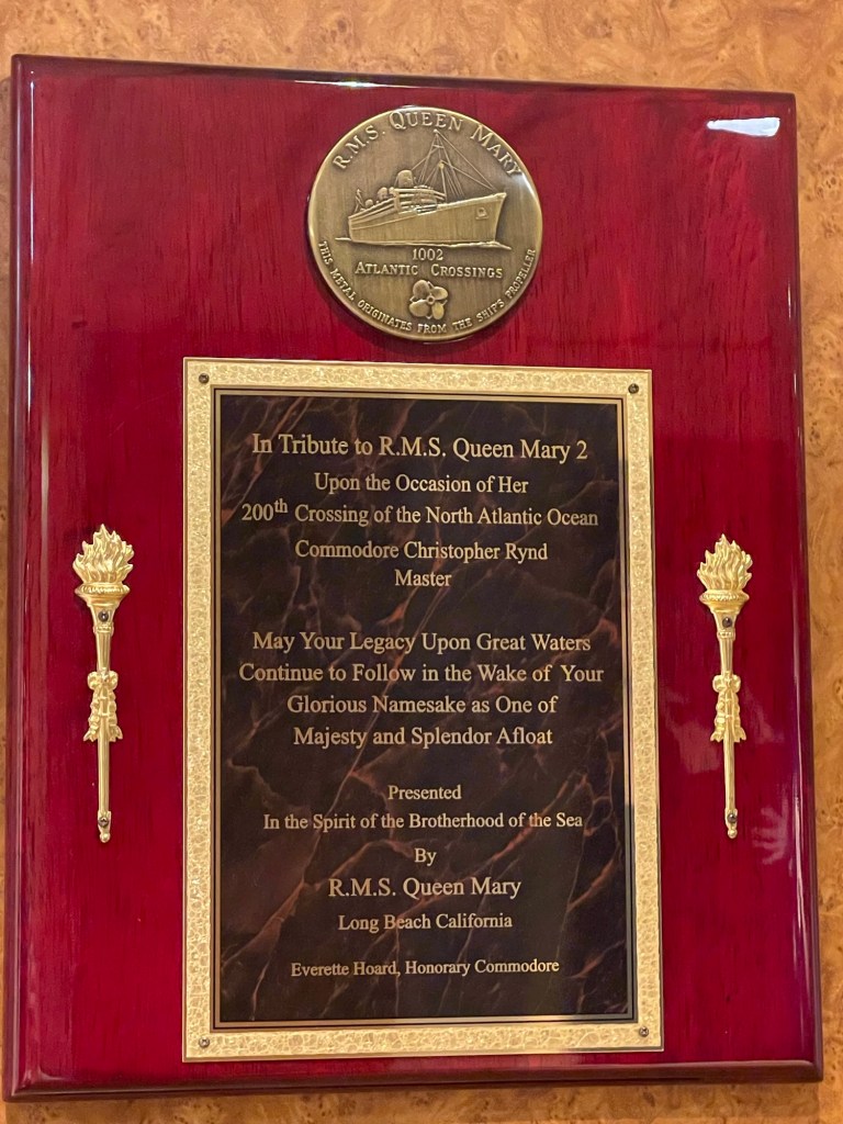 Wood plaque with gold embellishments, with black marble that reads In Tribute to R.M.S. Queen Mary 2 Upon the Occasion of Her 200th Crossing of the North Atlantic Ocean Commodore Christopher Rynd Master
May Your Legacy Upon Great Waters Continue to Follow in the Wake of Your Glorious Namesake as One of Majesty and Splendor Afloat Presented In the Spirit of the Brotherhood of the Sea By R.M.S. Queen Mary
Long Beach California Everette Hoard, Honorary Commodore. Round bronze piece has image of the liner Queen Mary and reads 1002 Atlantic crossings this medal originates from the ships master. 