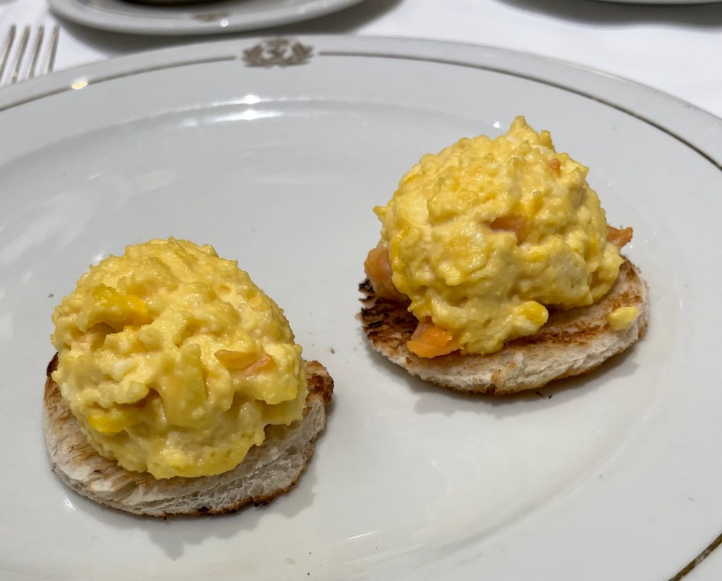Two thin wheat biscuits, each topped with a slice of smoked salmon and a scoop of fluffy scrambled eggs on white china bearing the Cunard emblem. 