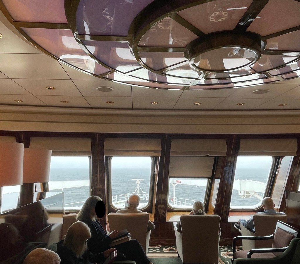 Passengers sit in individual chairs reading while overlooking the bow and the calm Atlantic Ocean on an overcast day. 