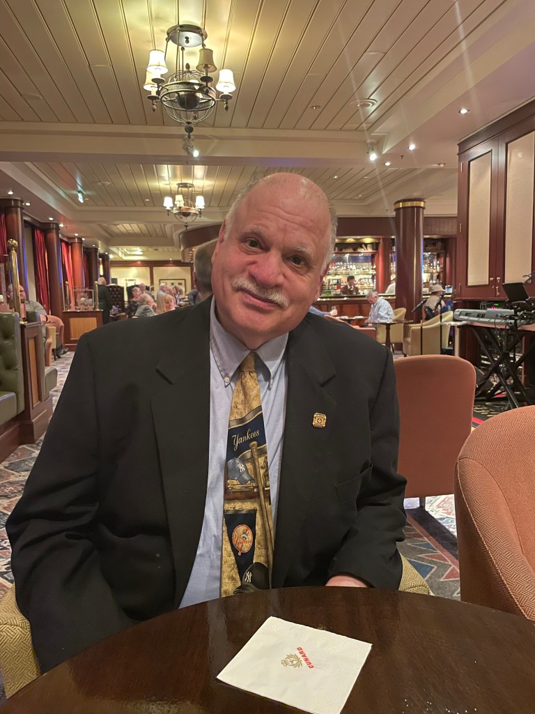 60 ish year old man in dark blazer, light shirt and New York Yankees tie sits at a round cocktail table in a bar with dark wood like columns, and wood wall trim. 