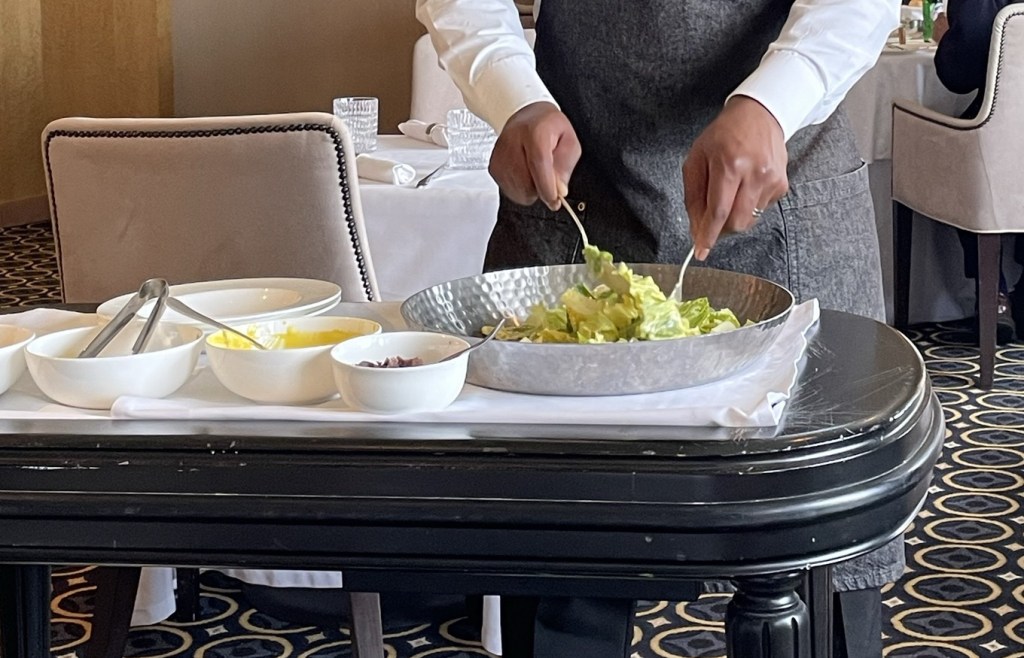 The hands of a formally dressed chef toss romaine lettuce in a wooden bowl atop a cart that also includes small bowls of cheese, croutons, and a yellow dressing