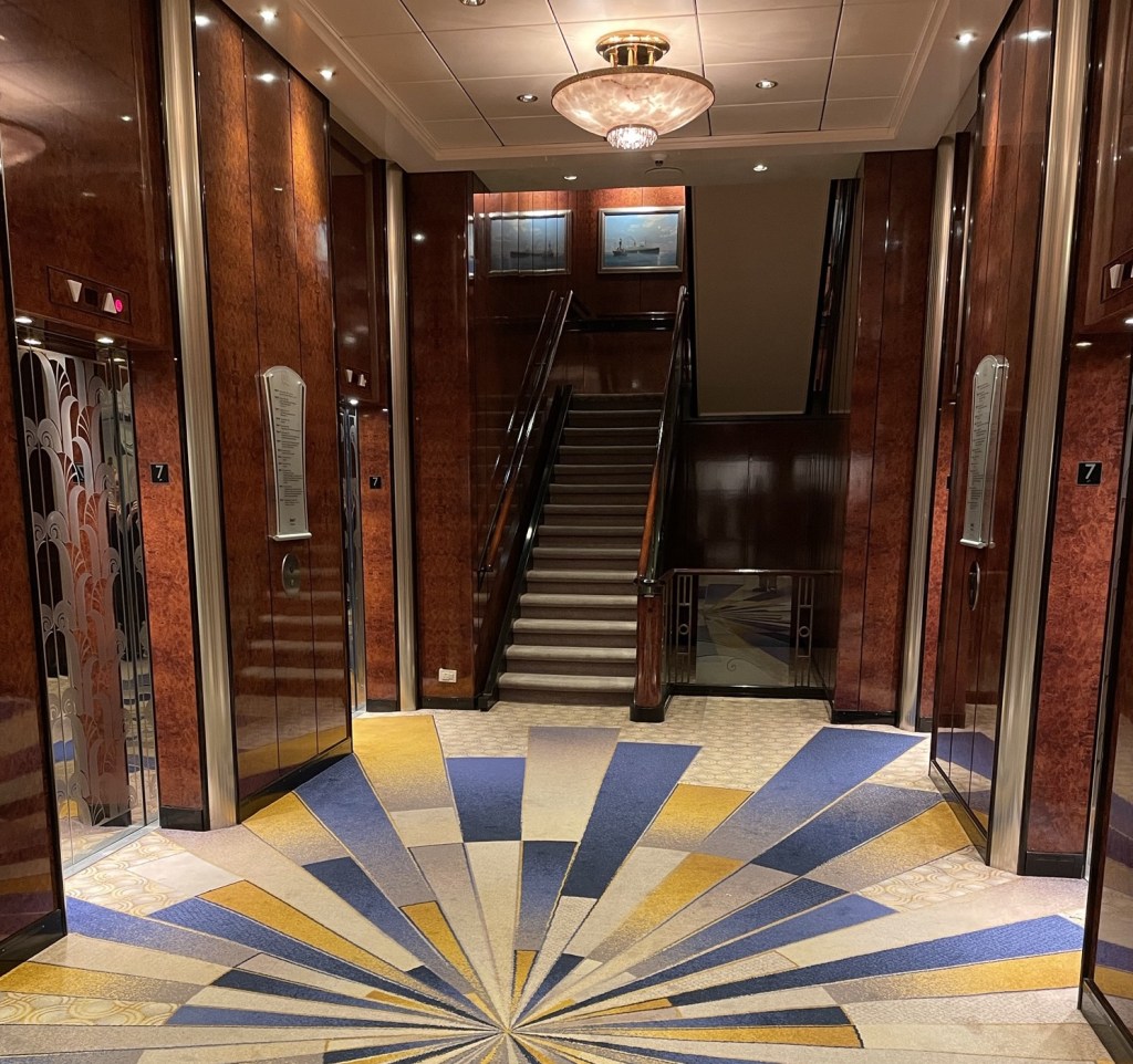 A starburst rug is in the elevator landing on a ships interior deck, with a staircase and ship artwork in the background. 