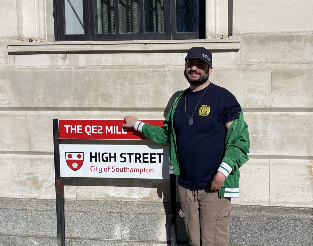 A young Caucasian man with dark beard and ball cap stands next to a sign marking the Q E 2 mile on High Street city of Southampton