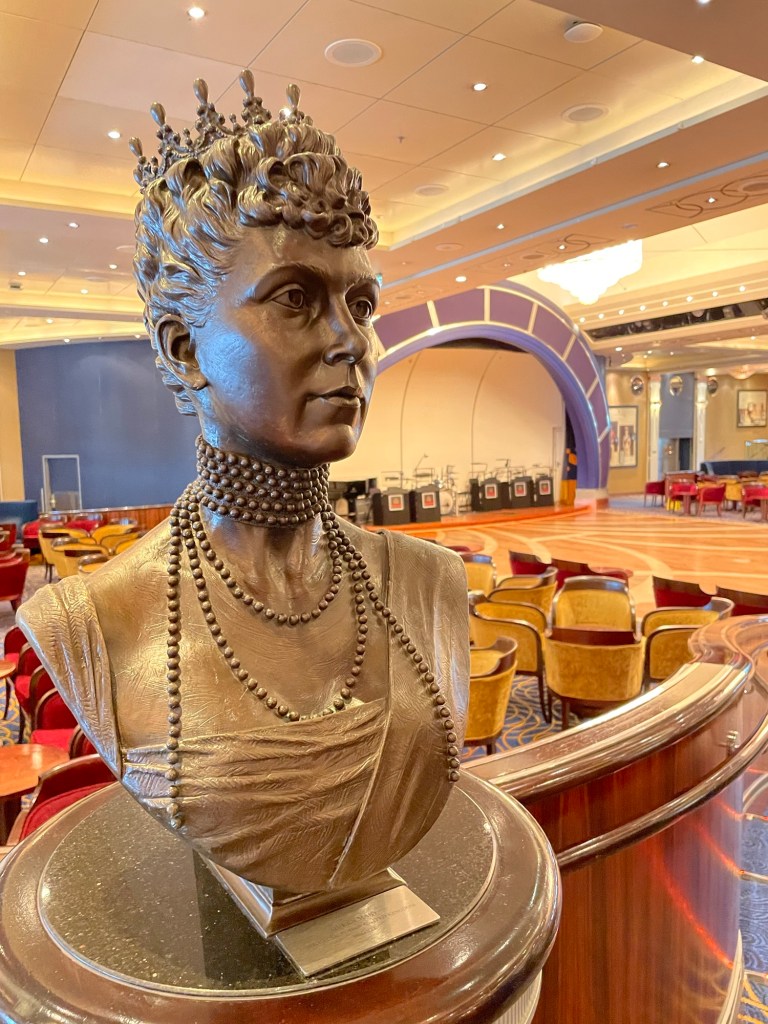 Bronze bust of Queen Mary wearing a beaded necklace and crown, displayed on a polished stand in an elegant lounge with a wood dance floor, plush seating, and a stage with bandstands.  