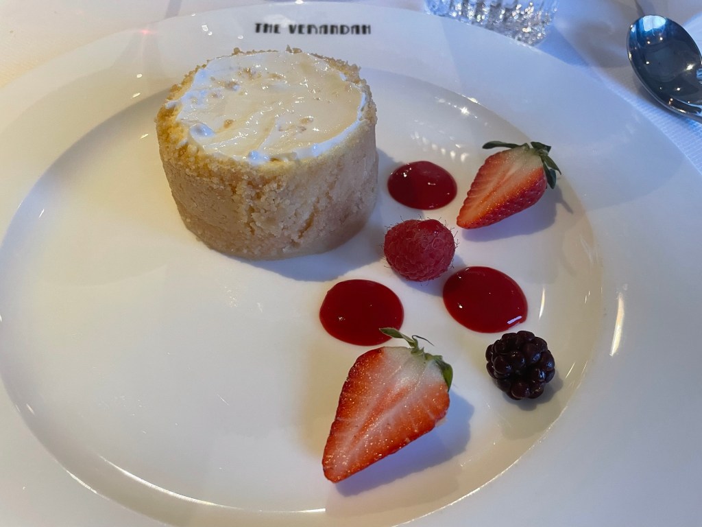 On a dessert plate a cylindrical crust filled with smooth cheesecake, and on the side two strawberry halves, a red raspberry a black raspberry and three dollops of red sauce