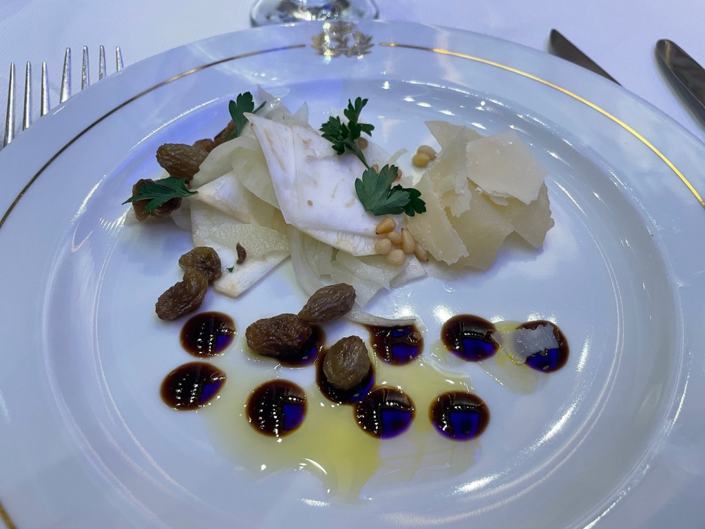 A round white dinner plate with Cunard logo on top rim shows the shaved pale yellow vegetable in the top half drizzled with raisins and celery root and garnished with parsley, and bottom half of plate has dolloped dressing and raisins. 