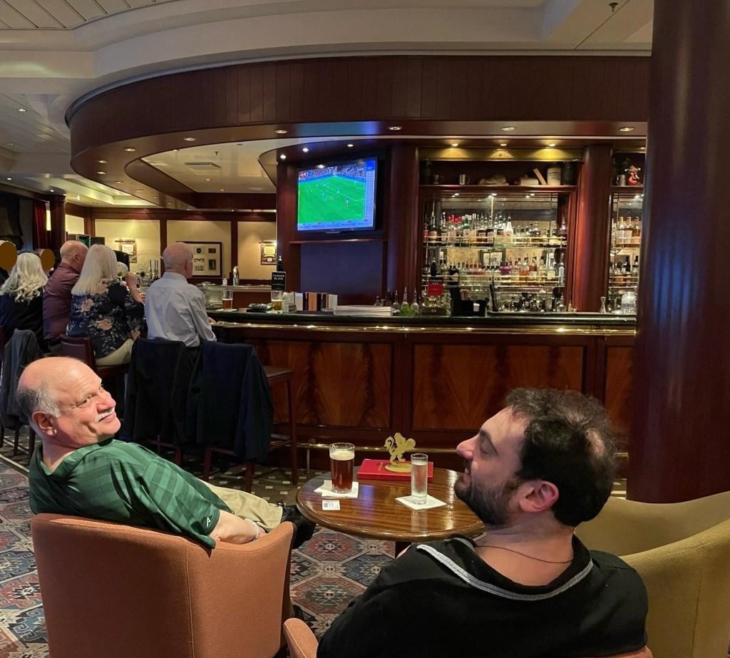 Neil and Marty turn their heads around in chairs that face a round dark wood bar with a TV showing a soccer foot ball game. Other patrons sit around the bar. 