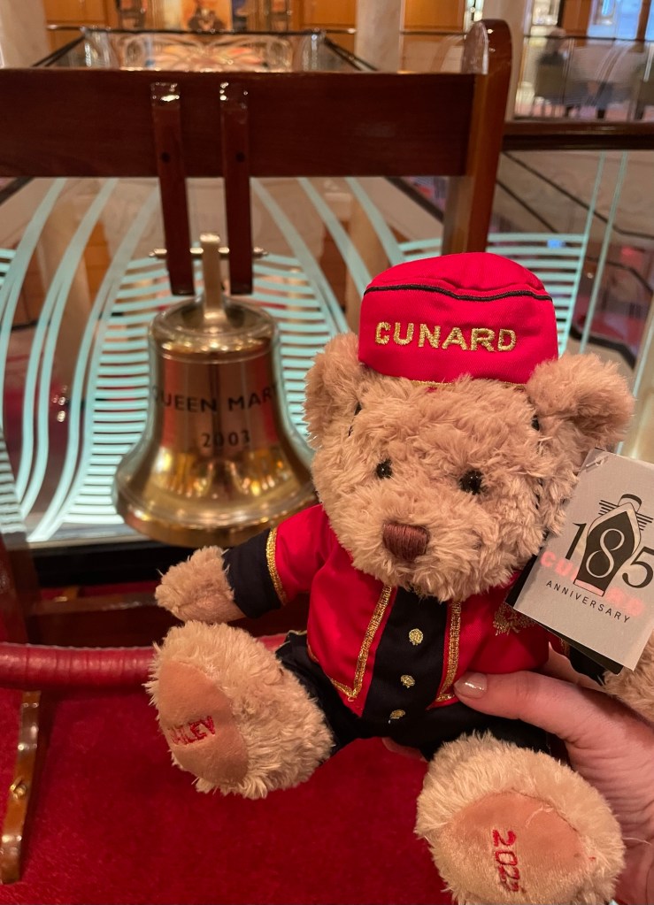 A plush teddy bear about 9 inches high dressed in a red Cunard bellhop uniform and cap is held in front of a ship’s bell engraved Queen Mary 2, 2003. The bear has 2025 stitched on one paw, the name Bailey on the other paw, and a tag reading “m185th Anniversary.