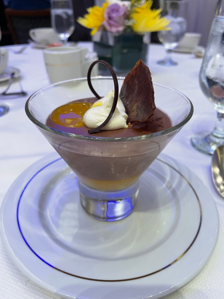 A chocolate mousse dessert in a v shaped glass, topped with shaved chocolate and a dollop of whipped cream 