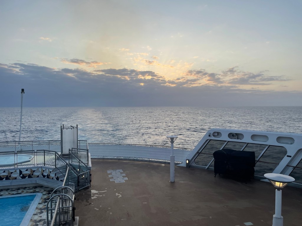 The sun disappears behind gray streaky clouds as viewed from the aft teak deck of a passenger ship. There’s a swimming pool on deck and the view of the wake is unobstructed. 