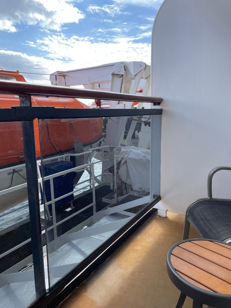 An orange topped lifeboat with white hull hangs on a davit alongside a glass balcony of a cruise ship cabin under a daytime sky with wispy clouds. 