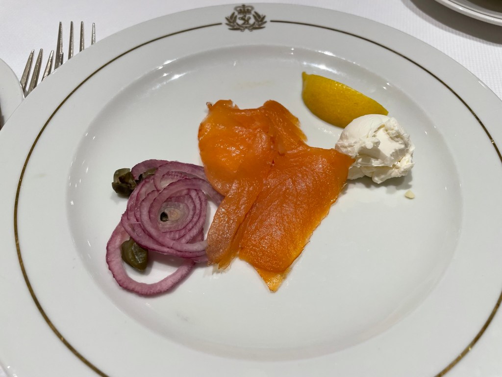 Red onion slices, capers, thinly sliced smoked salmon, scoop of cream cheese, and a lemon wedge on a gold rimmed plate