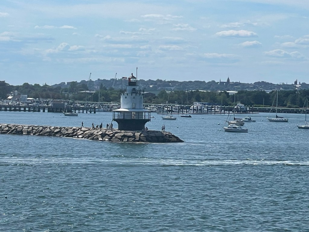 The spool like lighthouse with wraparound deck at bottom is positioned on a rocky jetty that juts out from the land. 